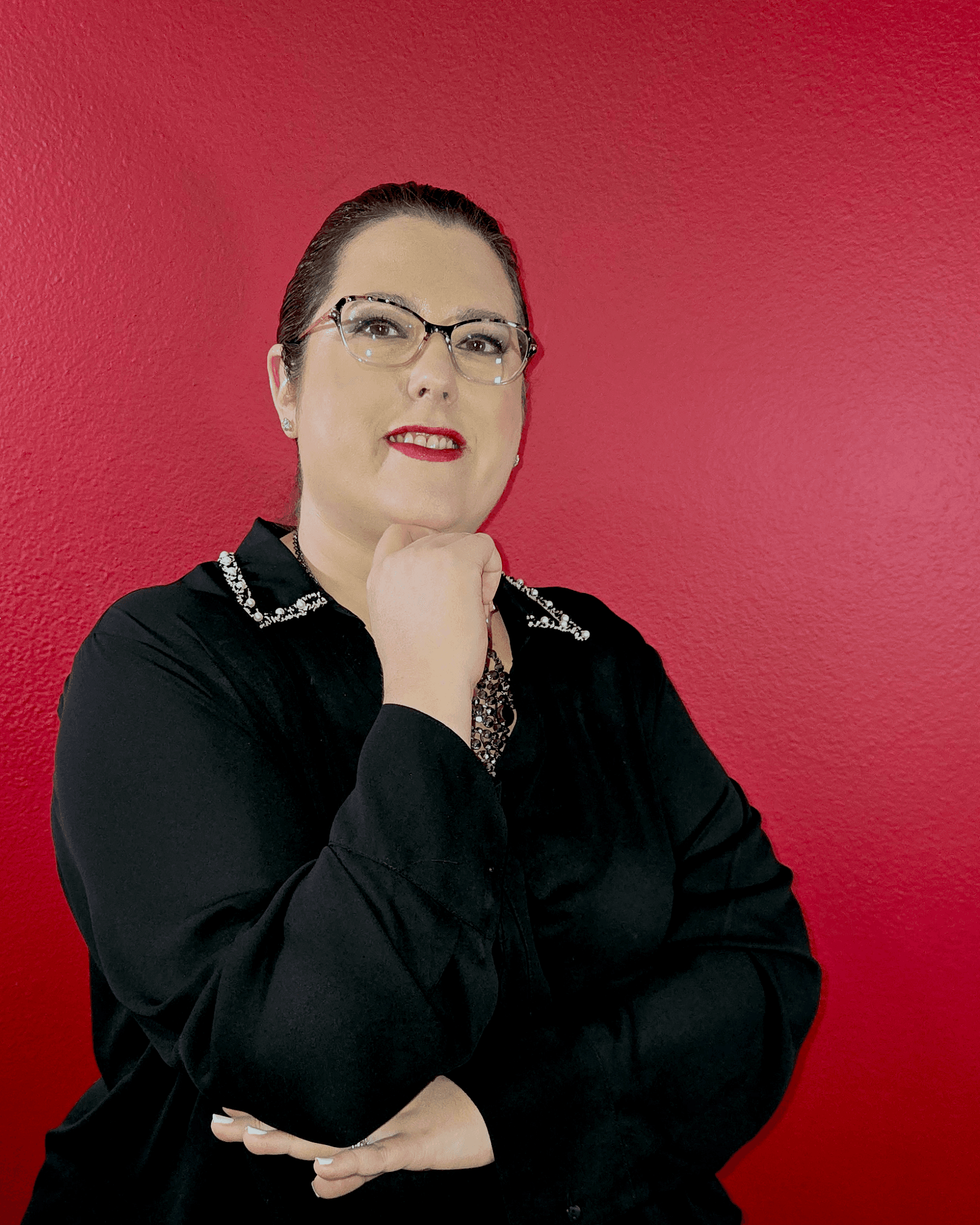 Woman with glasses and red lipstick poses with hand under chin against a red wall.